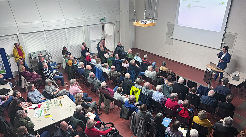 Eine Präsentation in einem Seminarraum mit vielen Teilnehmern, die auf Stühlen sitzen und einem Redner zuhören, der an einem Pult steht und eine Präsentationsfolie zeigt.